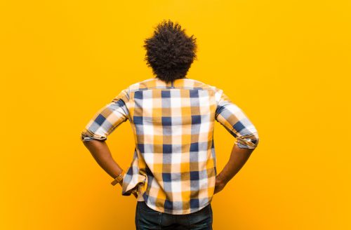man in a plaid shirt standing facing a yellow wall, with hands on his hips, looking confused or pensive