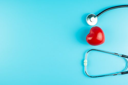 Top view of a stethoscope surrounding a red heart stress ball on a blue background.